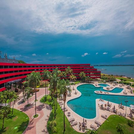 Royal Tulip Brasília Alvorada Hotel Exterior foto The photo shows a modern hotel building with a striking red exterior, surrounded by lush greenery. In the foreground, there is a large, creatively shaped swimming pool, with lounge chairs arranged around it. The backdrop features a body of water, pos